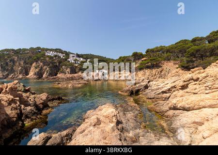 Le tranquille acque turchesi si infrangono dolcemente contro una costa rocciosa, con case bianche annidate su una scogliera sotto un cielo blu limpido in spagna Foto Stock