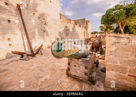 Antico cannone in bronzo su una carrozza in legno all'interno delle mura di un castello medievale affacciato sul mare, con ancora arrugginite e palle di cannone Foto Stock