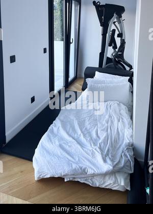 Il letto con biancheria bianca è posizionato in un angolo accogliente, accanto a una moderna macchina per esercizi, che unisce comfort e fitness in un ambiente elegante Foto Stock