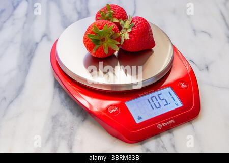Fragole fresche su un set rosso brillante di bilance digitali da cucina su un piano in marmo Foto Stock
