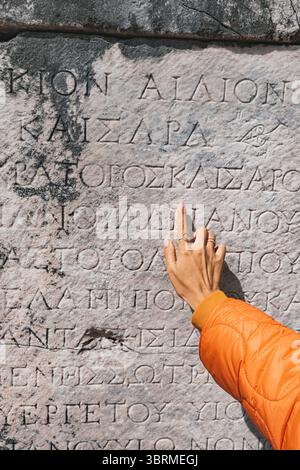 Il turista punta il dito contro l'antica iscrizione greca scolpita su una pietra di marmo Foto Stock