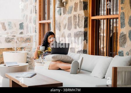 Una donna che si rilassa su un divano del patio, indossa un abito floreale e tiene in mano una tazza, mentre lavora su un notebook di fronte a un muro rustico in pietra Foto Stock