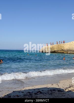 Le persone si sono riunite su una spiaggia per nuotare e provare l'emozione di saltare da una scogliera nelle acque cristalline in una giornata di sole. La scena cattura il Foto Stock