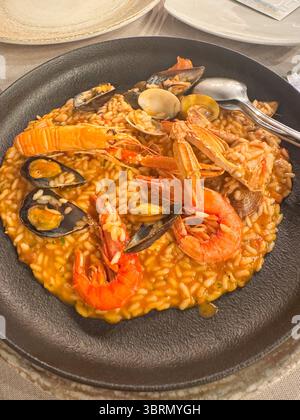 Delizioso risotto di pesce con gamberi, cozze e vongole servito su un piatto nero con forchetta e cucchiaio. Il piatto poggia su un tavolo, pronto Foto Stock
