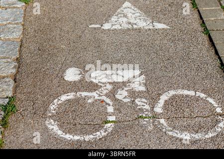 Primo piano tedesco di una pista ciclabile in asfalto usurata con simbolo della bicicletta dipinto di bianco e freccia direzionale. Bordo in pietra grigio visibile su entrambi i lati. Foto Stock