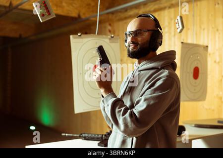 Un cliente del poligono di tiro arabo che ha in mano un'arma da fuoco e si gode un'esperienza ricreativa al chiuso. Uomo mediorientale che indossa indumenti protettivi che pratica con le armi in un ambiente sicuro e accogliente Foto Stock