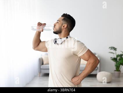 Giovane uomo arabo sportivo con cuffie bere acqua dopo il suo allenamento a casa Foto Stock