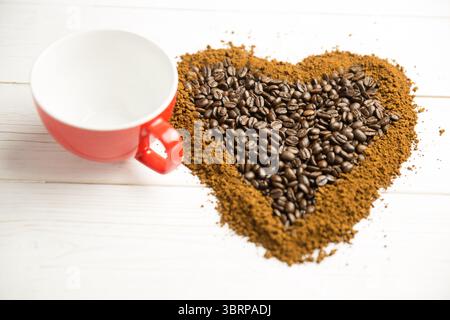 I fondi e i chicchi di caffè a forma di cuore si formano su una superficie piana bianca con una tazza rossa Foto Stock