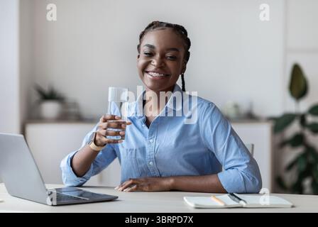 Idratazione al lavoro. Happy Black femmina lavoratore d'ufficio che tiene vetro d'acqua Foto Stock