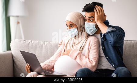 Scossa la coppia musulmana incinta in maschere mediche seduti con il laptop A casa Foto Stock