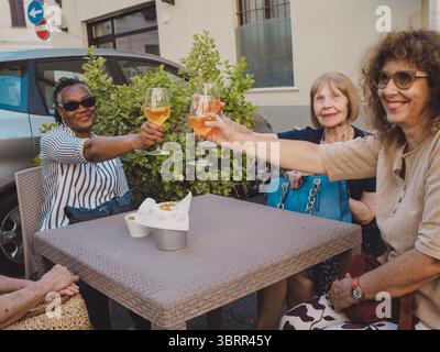 Un gruppo di donne anziane multi etniche che brindano con bicchieri di vino mentre si siede insieme a un tavolo all'aperto, godendosi un momento allegro di venerdì Foto Stock