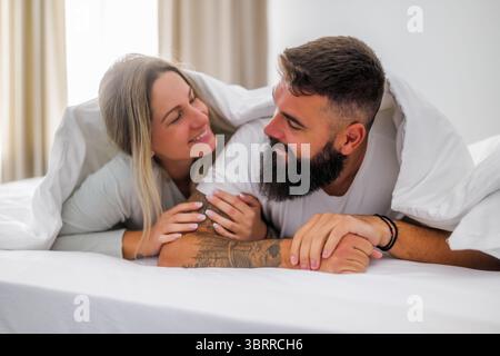 La mattina, una giovane coppia felice e amorevole è sdraiata a letto. Si stanno divertendo e sorridendo. Foto Stock