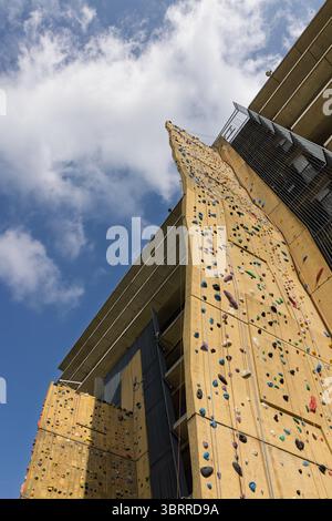 Una torreggiante parete per arrampicata all'aperto, piena di stive colorate, raggiunge un cielo blu luminoso con nuvole soffici, invitante avventura e sfida Foto Stock