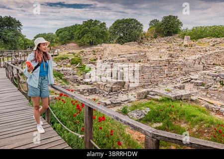 Una giovane donna che utilizza uno smartphone cammina su un passaggio pedonale in legno e visita le antiche rovine di Troia, un famoso sito archeologico in Turchia Foto Stock