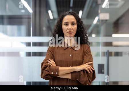 Una donna d'affari seria si trova di fronte a un edificio di uffici, con le braccia incrociate, guardando la telecamera. Foto Stock