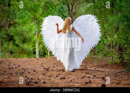Una splendida giovane bionda vestita di bianco puro, adornata da delicate ali d'angelo, crea una posa graziosa. La sua espressione serena e il suo abbigliamento fluido ev Foto Stock