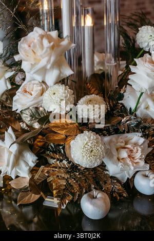 Un sofisticato allestimento floreale con rose bianche e dalie con foglie dorate e candele, che creano un caldo ed elegante centro per le feste Foto Stock