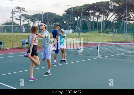 Amici diversi che si allungano su un campo da tennis all'aperto con racchette e rete da tennis visibile Foto Stock