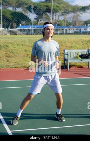 Tennista adolescente asiatico in piedi sul campo con fascia con panca, borsa e bottiglia d'acqua Foto Stock