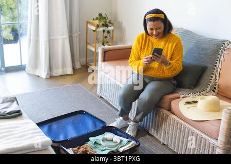 Donna rilassante che indossa un maglione giallo sul divano che controlla lo smartphone vicino a valigia aperta e spazio per le copie Foto Stock