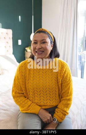 Donna seduta sul bordo del letto sorridente alla macchina fotografica in una camera luminosa con testiera e lampada capovolti Foto Stock
