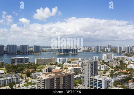 Vista aerea degli edifici che riflettono l'acqua tranquilla, con lo skyline che si estende sotto un cielo blu luminoso con nuvole soffici, Sunny Isles Beach, Florida, Stati Uniti. Foto Stock
