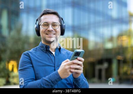 Un uomo che indossa cuffie e occhiali sorride mentre usa il telefono all'aperto, con un edificio sullo sfondo. Foto Stock