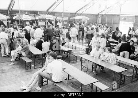 Settimana di festa Heemskerk parti Heemskerk, Whizgle News, Dutch Desk, Paesi Bassi, 1950 - 2000 il 04-09-1985. L'immagine contiene questi argomenti. La scena cattura un vivace raduno al chiuso, probabilmente ambientato in una grande struttura simile a una tenda con un'atmosfera temporanea o festosa. L'atmosfera è piena di un mix di eccitazione e interazione sociale, caratterizzata da gruppi di persone impegnate nella conversazione. In primo piano, diverse panchine e tavoli in legno, leggermente intemprati, sono disposti in modo un po' informale e invitante. Gruppi di persone occupano queste panchine, alcuni seduti cl Foto Stock