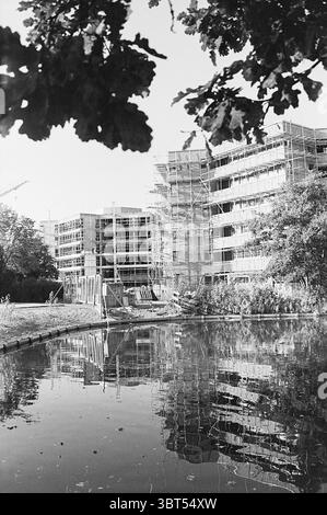 Relazione sulla nuova costruzione in Heemstede relazione, Whizgle News, Dutch Desk, Paesi Bassi, 1950 - 2000 il 08-10-1970. L'immagine mostra questi argomenti. La scena cattura un cantiere che presenta più edifici in fase di sviluppo, incorniciati dalla serena presenza di acqua. Il primo piano è costituito da un corpo d'acqua calmo che rispecchia perfettamente le strutture che si innalzano dietro di esso, creando un riflesso armonioso. Gli edifici sono parzialmente oscurati da impalcature, con telai in legno e materiali esterni visibilmente in fase di applicazione. Intorno all'area di costruzione, sfumature di gr Foto Stock