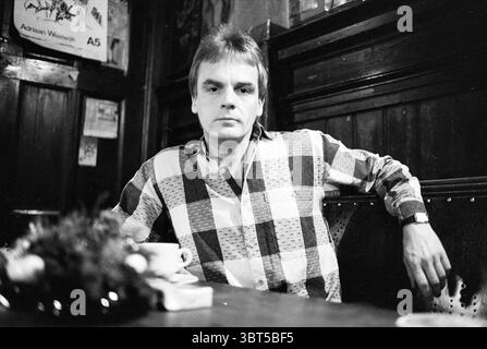 Rob Elson organizzatore, tra le altre cose, dell'organizzazione di Amsterdam per le elezioni dei bambini. Ufficio per l'affitto della camera persone Amsterdam Paesi Bassi, Whizgle News, Dutch Desk, Paesi Bassi, 1950 - 2000 il 30-12-1986. Questi sono gli argomenti nell'immagine. In un ambiente scarsamente illuminato, un uomo siede con fiducia su un tavolo di legno. Indossa una camicia a scacchi con motivi audaci in colori contrastanti, che cattura l'attenzione dell'ambiente rustico. Il design della camicia presenta diverse tonalità di luce e di buio, creando una profondità visiva che si integra perfettamente con l'ambiente circostante. La sua espressione è premurosa, forse respinta Foto Stock