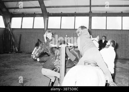 Festeggiamenti Spaarndam Overveen Rozenprieel Asiaweg Queen's Day Haarlem Aziëweg Paesi Bassi, Whizgle News, Dutch Desk, Paesi Bassi, 1950 - 2000 il 30-04-1974. Questi sono gli elementi dell'immagine. In una spaziosa arena interna con un alto soffitto in legno, l'atmosfera è piena di emozioni ed energia, mentre un gruppo di bambini interagisce con due cavalli. L'ambiente è caldo e invitante, con luce naturale che scorre attraverso grandi finestre che si estendono su un lato, proiettando ombre morbide sul pavimento di sabbia. Una ragazza con un cappotto scuro si aggrappa alla schiena di un piccolo cavallo marrone, la sua coscia Foto Stock