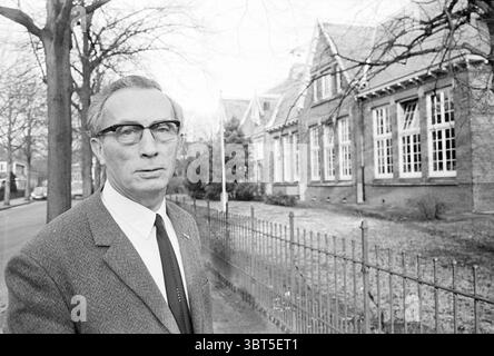 Ritratto del capo della St Joseph School Schools, Whizgle News, Dutch Desk, Paesi Bassi, 1950 - 2000 il 19-11-1971. L'immagine contiene questi argomenti. In un ambiente urbano sereno, un uomo distinto si trova in primo piano, trasudando un senso di quiete autorità. Indossa un abito sartoriale grigio scuro impreziosito da una camicia bianca nitida e una cravatta stretta, il tutto per aggiungere un aspetto sofisticato. La sua acconciatura è ben pettinata e sfoggia occhiali sottili e rettangolari che sottolineano il suo comportamento premuroso. Dietro di lui, un edificio in mattoni presenta grandi finestre a più pannelli, che riflettono Foto Stock