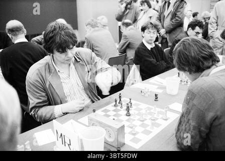 Municipal Chess Council IJmond Trophy Blast Furnace Chess Tournament, Whizgle News, Dutch Desk, Paesi Bassi, 1950 - 2000 il 15-01-1989. Questi sono gli elementi dell'immagine. In un vivace ambiente interno, la scena è dominata da un lungo tavolo ricoperto da un panno di feltro verde, su cui è esposta una scacchiera. I pezzi degli scacchi sono disposti a metà partita, catturando la tensione del pensiero strategico. Seduti al tavolo, due giocatori sono impegnati in una profonda concentrazione. Un giocatore, con i capelli scuri e vestiti con un blazer beige casual, si inclina in avanti, con le dita delicate Foto Stock