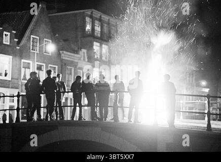 Falò sul ponte Burgwal durante Capodanno Haarlem Burgwal Paesi Bassi, Whizgle News, Dutch Desk, Paesi Bassi, 1950 - 2000 il 31-12-1966. Questi sono gli argomenti nell'immagine. In un ambiente notturno, un gruppo di persone si staglia di fronte a un'esplosione luminosa e ardente. L'illuminazione proietta ombre spettacolari, sottolineando le figure come forme scure sullo sfondo illuminato. Scintille e brace ballano verso l'alto dalla sorgente luminosa, creando un contrasto dinamico con l'oscurità che la circonda. Gli individui sono riuniti su un ponte, le loro posture suggeriscono un mix di fascinatio Foto Stock