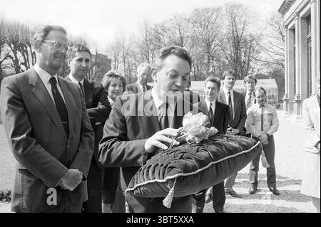 Presentazione del Ministro europeo dei tulipani Braks Flowers and Flower Girls presentazione, Whizgle News, Dutch Desk, Paesi Bassi, 1950 - 2000 il 14-04-1988. Questi sono gli elementi dell'immagine. Un gruppo di individui è radunato all'aperto, apparentemente in un ambiente formale o significativo. La scena ha un'aura storica o cerimoniale, suggerita dall'elegante architettura sullo sfondo, che sembra essere un grande edificio con grandi finestre e un design classico. In primo piano, un uomo vestito con un abito affilato regge un grande cuscino scuro ornato da motivi intricati. Sta posizionando con attenzione o. Foto Stock