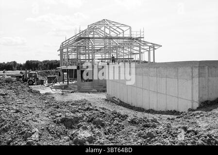Telaio in acciaio di un capannone, Whizgle News, Dutch Desk, Paesi Bassi, 1950 - 2000. Questi sono gli argomenti nell'immagine. La scena raffigura un cantiere in corso sotto un cielo limpido. La struttura in costruzione presenta una struttura scheletrica di legno che si erge in modo prominente, mostrando il suo stato incompiuto con travi che sporgono e un tetto angolato che prende forma. La combinazione di elementi naturali e artificiali crea un senso di operosità. Intorno all'edificio c'è un fresco strato di terra e ghiaia, che indica il recente movimento terra, che dona all'area un tono grezzo e terroso. Un grande foun di cemento Foto Stock