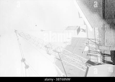 Transport Vault Bank Transport, Whizgle News, Dutch Desk, Paesi Bassi, 1950 - 2000 on 26-11-1970. L'immagine contiene questi argomenti. La scena cattura un cantiere, caratterizzato dalla presenza di una gru alta, che si estende verso l'alto contro il cielo luminoso. L'intricata struttura metallica della gru spicca, gettando ombre lunghe e scure sulle superfici sottostanti. Intorno alla gru, gli edifici sono parzialmente visibili, mostrando varie consistenze di mattoni e cemento. Le sommità di queste strutture sono apparentemente illuminate dalla luce del sole, dando un netto contrasto con le più scure e ombreggiate Foto Stock