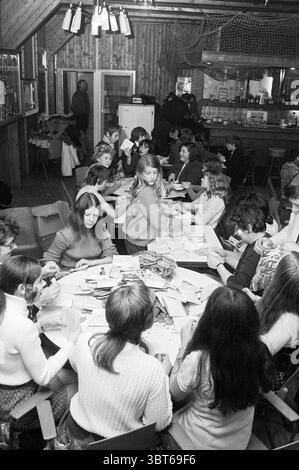 Tough Packing Beverwijk St. Nicholas Sinterklaas, Whizgle News, Dutch Desk, Paesi Bassi, 1950 - 2000 il 17-11-1971. L'immagine include questi argomenti. La scena si snoda all'interno di uno spazio rustico e caldo illuminato caratterizzato da pareti in legno e da un'atmosfera intima. Un lungo tavolo domina il centro, circondato da un mix di adulti e bambini, tutti impegnati in varie attività. Sembrano essere coinvolti in qualche forma di creazione o creazione, con una serie di materiali colorati sparsi su tutta la tavola: Carte, forbici e altri strumenti artistici. Gli individui esprimono una serie di espressioni, Foto Stock