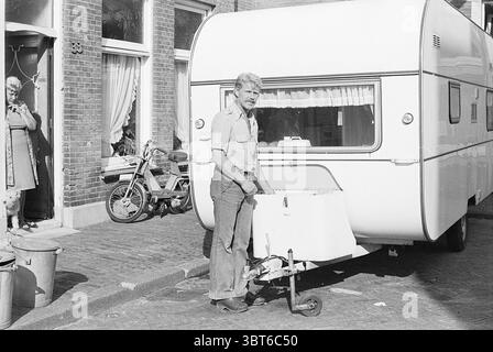 Sig. Goebert presso Caravan Persons, Whizgle News, Dutch Desk, Paesi Bassi, 1950 - 2000 il 03-08-1976. L'immagine contiene questi argomenti. La scena presenta un angolo di strada soleggiato con una carovana bianca e luminosa ben parcheggiata. La roulotte ha un esterno liscio e lucido e un design semplice e moderno con bordi rotondi, che creano un senso di apertura. Di fronte a essa, un uomo di mezza età sta in piedi, afferrando la maniglia di una piccola piattaforma a ruote collegata alla roulotte. Indossa una camicia a maniche corte di colore chiaro e appare concentrato, con un leggero solco sulla fronte. Dietro di lui, l'ambientazione è caratteristica Foto Stock