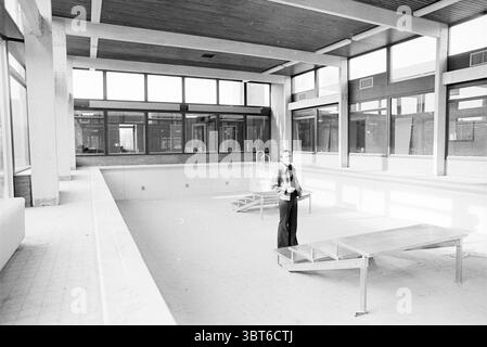 Ritrattista in piedi nella piscina coperta vuota con Foto interni, Whizgle News, Dutch Desk, Paesi Bassi, 1950 - 2000 nel 1977. Questi argomenti vengono visualizzati nell'immagine. La scena raffigura un'ampia area piscina interna priva di acqua, che presenta un'estetica inquietante ma minimalista. La piscina stessa è poco profonda e piena di una superficie chiara e aspra, che ne sottolinea il vuoto. Intorno alla piscina sono presenti grandi finestre che si estendono dal pavimento al soffitto, permettendo alla luce naturale di inondare lo spazio, creando un'atmosfera luminosa e ariosa. La luce proietta ombre morbide attraverso il ro Foto Stock