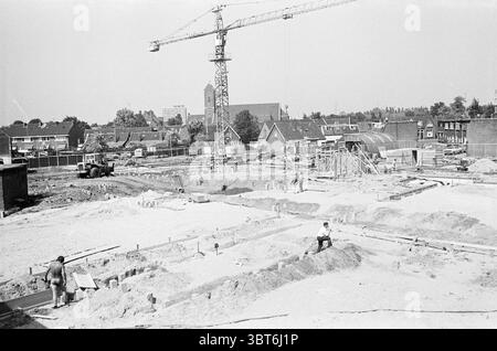 Panoramica del cantiere vicino alla biblioteca Beverwijk siti, Whizgle News, Dutch Desk, Paesi Bassi, 1950 - 2000 il 04-08-1975. Questi argomenti sono mostrati nell'immagine. La scena cattura un vivace cantiere sotto un cielo chiaro e limpido. La luce del sole proietta ombre forti, evidenziando le varie trame e colori dei materiali sparsi per il terreno. In primo piano, diversi lavoratori sono impegnati in compiti diversi; alcuni stanno scavando e livellando la terra sabbiosa, mentre altri stanno posizionando travi e strutture in legno per quella che sembra essere la base di un edificio. Il loro coagulo Foto Stock