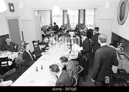 Ristorante economico Vincent Haarlem Nieuwe Groenmarkt Paesi Bassi, Whizgle News, Dutch Desk, Paesi Bassi, 1950 - 2000 il 13-07-1965. L'immagine contiene questi argomenti. In un vivace ambiente da pranzo, un lungo tavolo rettangolare si estende lungo la sala, circondato da un mix di commensali impegnati in conversazione e gustando i loro pasti. Il tavolo è allestito con semplici stoviglie e posate bianche, mentre alcuni piatti contengono ancora i resti di cibo. I partecipanti sono un gruppo eterogeneo, con uomini e donne, vestiti con una varietà di abiti tipici di un'epoca passata, alcuni in abiti e cravatte, altri in abiti e altro ancora Foto Stock