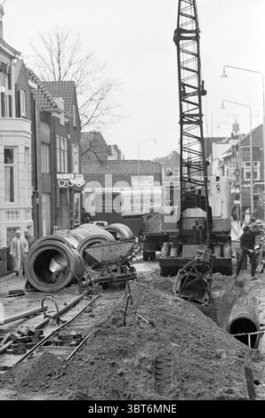 Lavori di scavo per fognature nello Zeestraat a Beverwijk Beverwijk Zeestraat Paesi Bassi ., Whizgle News, Dutch Desk, Paesi Bassi, 1950 - 2000. L'immagine contiene questi argomenti. La scena raffigura una strada in costruzione, creando un'atmosfera un po' caotica ma industriosa. In prima linea, diversi tubi cilindrici in cemento sono disposti sui lati, a indicare il lavoro in corso sotto la superficie. Intorno a loro c'è un pezzo di terra marrone scuro, che indica i recenti scavi. A destra si solleva una gru alta, la sua struttura metallica contrasta nettamente con la più morbida Foto Stock