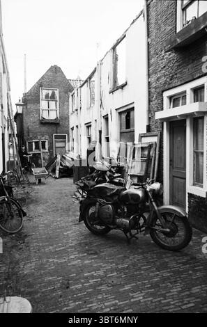 Strada senza uscita con tre uomini davanti a un edificio di demolizione, Whizgle News, Dutch Desk, Paesi Bassi, 1950 - 2000. Questi sono gli argomenti nell'immagine. La scena cattura uno stretto vicolo acciottolato fiancheggiato da edifici intemprati, le loro facciate bianche sbucciate rivelano l'età e l'abbandono. Un edificio si erge parzialmente rovinato, la sua struttura in legno esposta, mentre un altro presenta finestre con pannelli rotti, aggiungendo un senso di abbandono. Tra i detriti, una motocicletta d'epoca si trova in posizione dominante, con la sua superficie metallica che mostra segni di usura e ruggine, posizionata su ciottoli irregolari. Intorno a questo Foto Stock