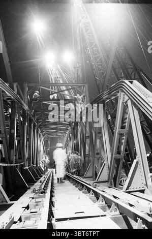 Sostituire il ponte ferroviario, Whizgle News, Dutch Desk, Paesi Bassi, 1950 - 2000 il 14-05-1992. L'immagine mostra questi argomenti. La scena cattura un grande cantiere illuminato da luci luminose che proiettano un forte bagliore, creando un contrasto drammatico con l'ambiente circostante più scuro. Ponteggi e travi in acciaio dominano il telaio, mostrando complessi design industriali con angoli e linee nitide che trasmettono un senso di precisione ingegneristica. Le superfici sono un mix di trame di metallo grezzo, alcune lucide e riflettenti, mentre altre sono grintose e intemperie. In mezzo a questo ambiente Foto Stock