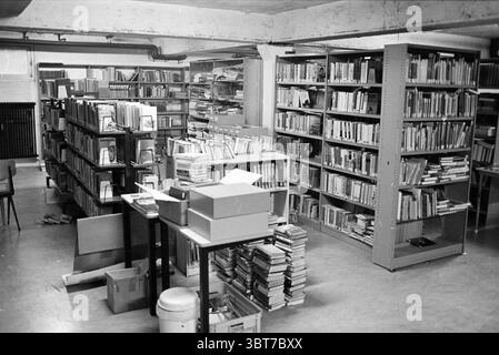 Interno della biblioteca, Whizgle News, Dutch Desk, Paesi Bassi, 1950 - 2000 il 10-1982. L'immagine mostra questi argomenti. La scena presenta un seminterrato poco illuminato che assomiglia a una biblioteca o a un archivio, pieno di un assortimento di libri e materiali. L'atmosfera trasmette un senso di tranquilla solitudine, invitante concentrazione e riflessione. In primo piano, alcuni tavoli sono sparsi, ingombrati da documenti, documenti e un assortimento di scatole di cartone impilate in modo casuale. Alcune scatole sono aperte, rivelando il loro contenuto, mentre altre si siedono chiuse, aggiungendo una sensazione disorganizzata dello spazio. Un'immondizia Foto Stock