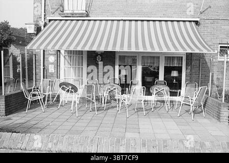 Terrace hotel Zandvoort, Whizgle News, Dutch Desk, Paesi Bassi, 1950 - 2000 il 09-1972. L'immagine contiene questi argomenti. La scena raffigura un modesto patio esterno, caratterizzato da una miscela di tessuto e metallo. A dominare l'area c'è una grande tenda a strisce, adornata da bande alternate di rosso e bianco, che offre ombra allo spazio sottostante. Il pavimento del patio e' pavimentato con piastrelle di cemento quadrato, che conferiscono un aspetto strutturato all'ambiente. In primo piano, diverse sedie in metallo bianco sono allineate in una disposizione un po' casuale, con le loro strutture sottili che contrastano con le più robuste Foto Stock
