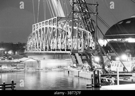 Sostituire il ponte ferroviario, Whizgle News, Dutch Desk, Paesi Bassi, 1950 - 2000 il 14-05-1992. Questi sono gli elementi dell'immagine. La scena è ambientata sul lungomare di notte, dove è in corso un grande progetto di costruzione. Un enorme ponte in acciaio viene sollevato in posizione da una gru torreggiante, il suo motivo geometrico di travi e capriate crea una silhouette impressionante contro il cielo scuro. L'acqua riflette le luci dell'area di lavoro, creando un effetto scintillante che contrasta con l'oscurità circostante. Varie barche sono legate lungo i moli, i cui scafi sono illuminati da morbidi e artificiali Foto Stock