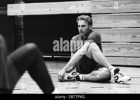 Foto allenamento Velser Basketball Club Akrides, Whizgle News, Dutch Desk, Paesi Bassi, 1950 - 2000 il 11-11-1992. L'immagine contiene questi argomenti. In una palestra, un uomo è seduto su un pavimento liscio di legno, trasuda un comportamento informale ma concentrato. Ha una struttura atletica, vestita con una semplice camicia grigia e pantaloncini neri, suggerendo di essere nel bel mezzo di un allenamento o di una sessione di allenamento. I suoi capelli sono biondi sabbiosi e leggermente duri, aggiungendo un aspetto rilassato. Lo sfondo presenta file di sbiancanti in legno lucido, che esaltano la sensazione di uno spazio sportivo ben utilizzato. Il li Foto Stock