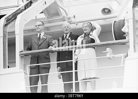 H.M. e il principe lasciano IJmuiden in barca ricevimenti reali e visite reali, Whizgle News, Dutch Desk, Paesi Bassi, 1950 - 2000 il 05-06-1975. Questi argomenti vengono visualizzati nell'immagine. La scena raffigura un trio di individui in piedi su un balcone o un ponte, probabilmente a bordo di una nave. La figura centrale, un uomo con capelli e occhiali a schiena affilati, è vestita con un abito scuro, che suggerisce un evento formale. Si trova tra due altri, tutti e tre mantengono una postura verticale che indica sicurezza e padronanza. Alla sua destra, una donna anziana con i capelli ricci, indossa un vestito di colore chiaro e una cardiga bianca Foto Stock